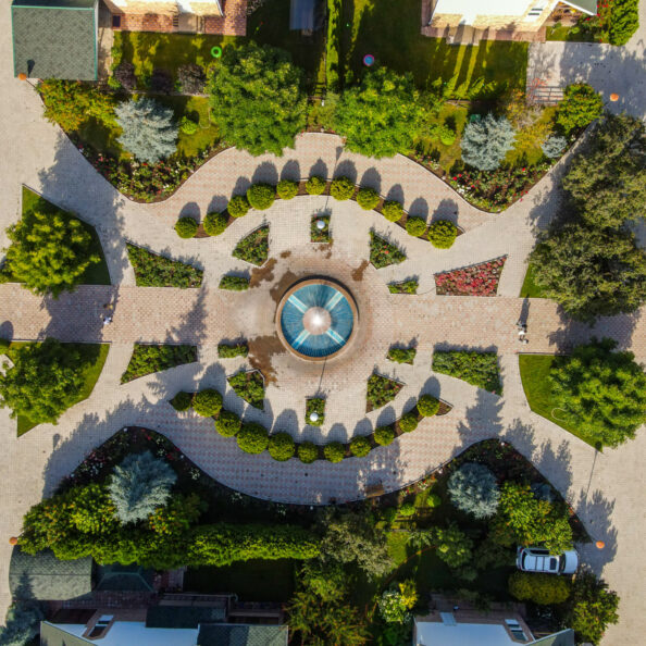 A top-down aerial view of a resort's decorative garden with a central fountain, hedges, flowers, and walkways
